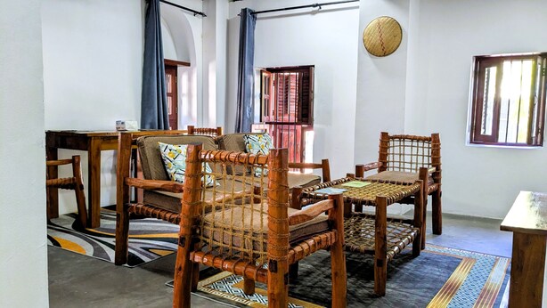 A room with a sofa, two wooden chairs, a small table, a patterned rug, and a window with curtains. - The Hub Train House Amazing Gents code (Zanzibar)