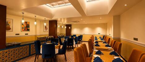 A hotel dining room with a long table set for a meal, multiple chairs, and a buffet area with food service equipment.