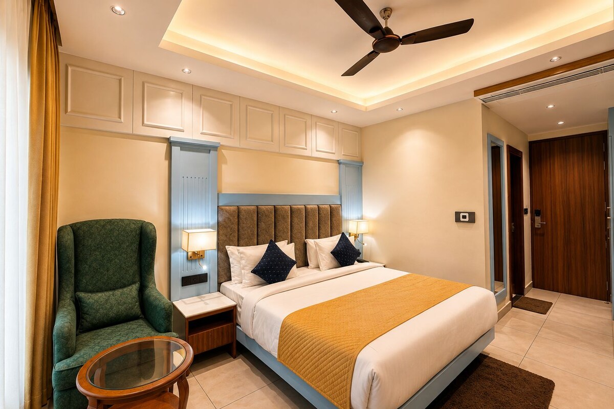 A hotel room with a bed, a green armchair, a small table, a ceiling fan, and a nightstand with a lamp.