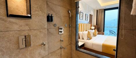A hotel bathroom with a large mirror, a shower, a sink, and a view of a balcony with a bed.
