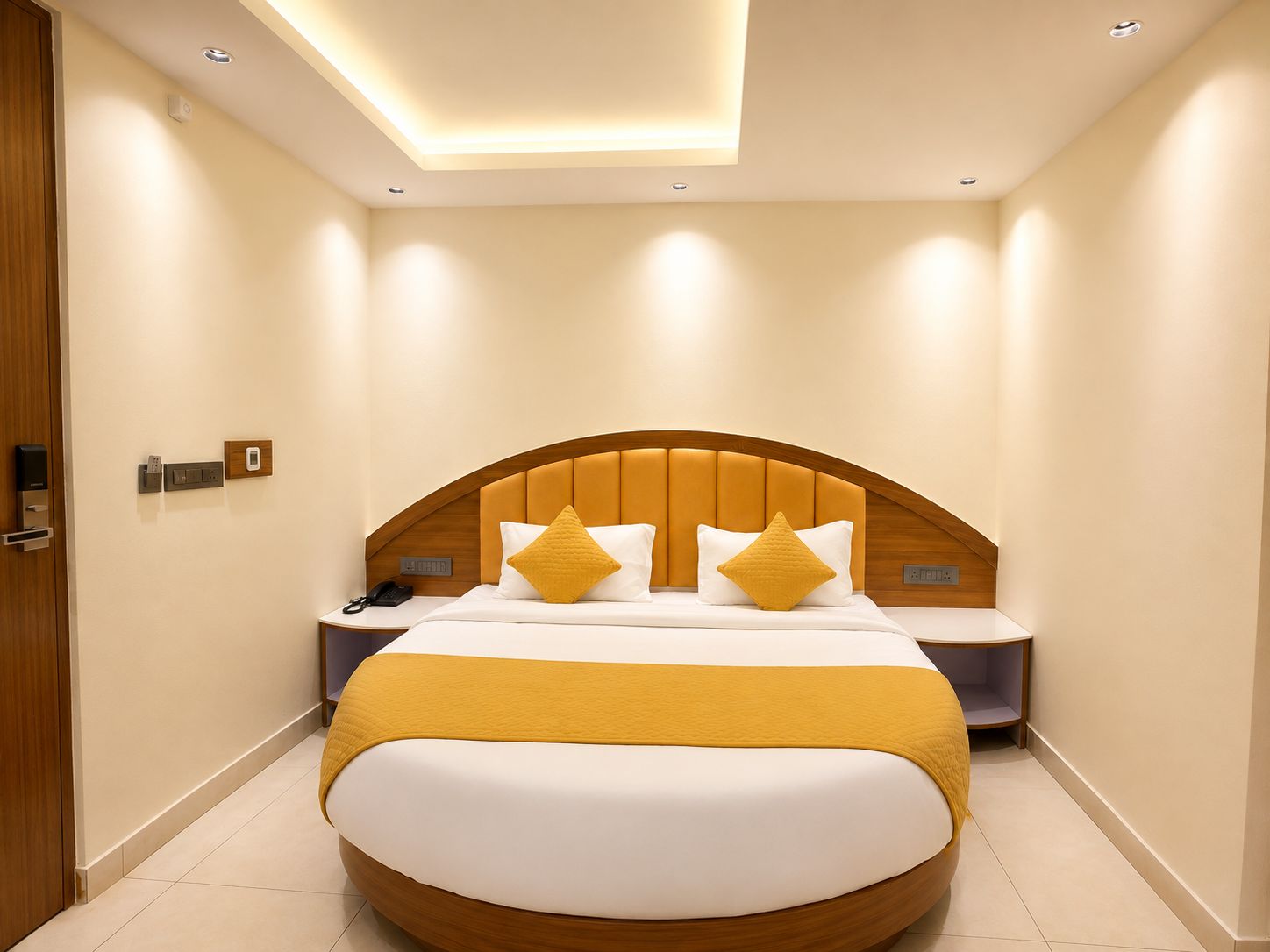 A modern hotel room with a curved bed, a wooden headboard, and a bedside table with a phone and a lamp.