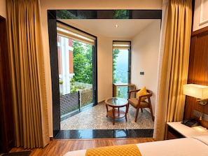A hotel room with a bed, a small table, a chair, a nightstand, and a view of the outside. - SSH Ardmore Hills (Mussoorie)