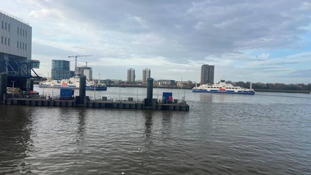 A waterfront dock with a ferry, a building with a crane, and several high-rise buildings in the background. - cosy 1bed perfect for family and contractors (Woolwich, London)