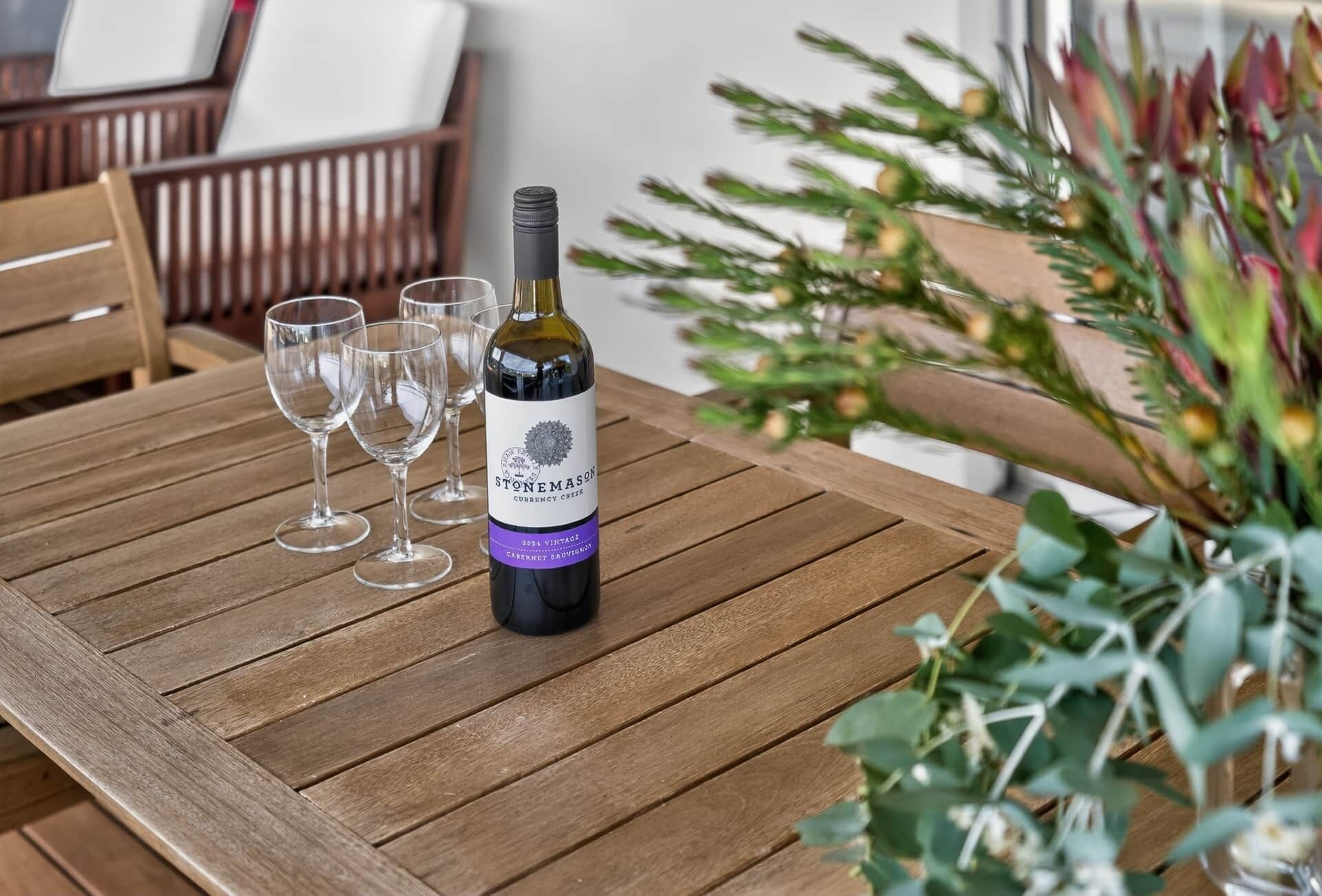 Una mesa de madera con una botella de vino Stonemason Currency Cuvée, cuatro copas de vino vacías y una planta en maceta.