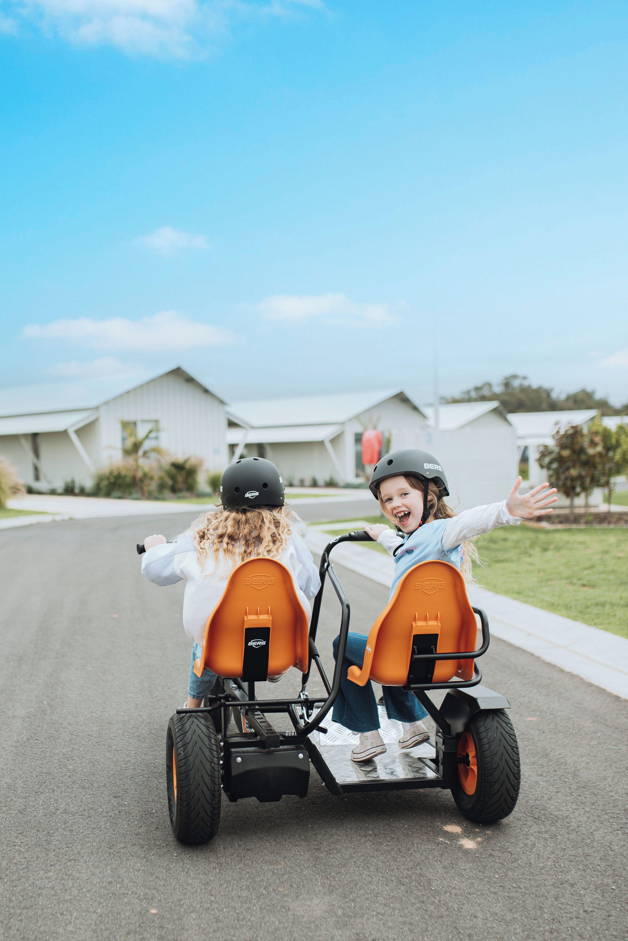 Two children on a stationary tricycle with a Berg logo, parked on a residential street.