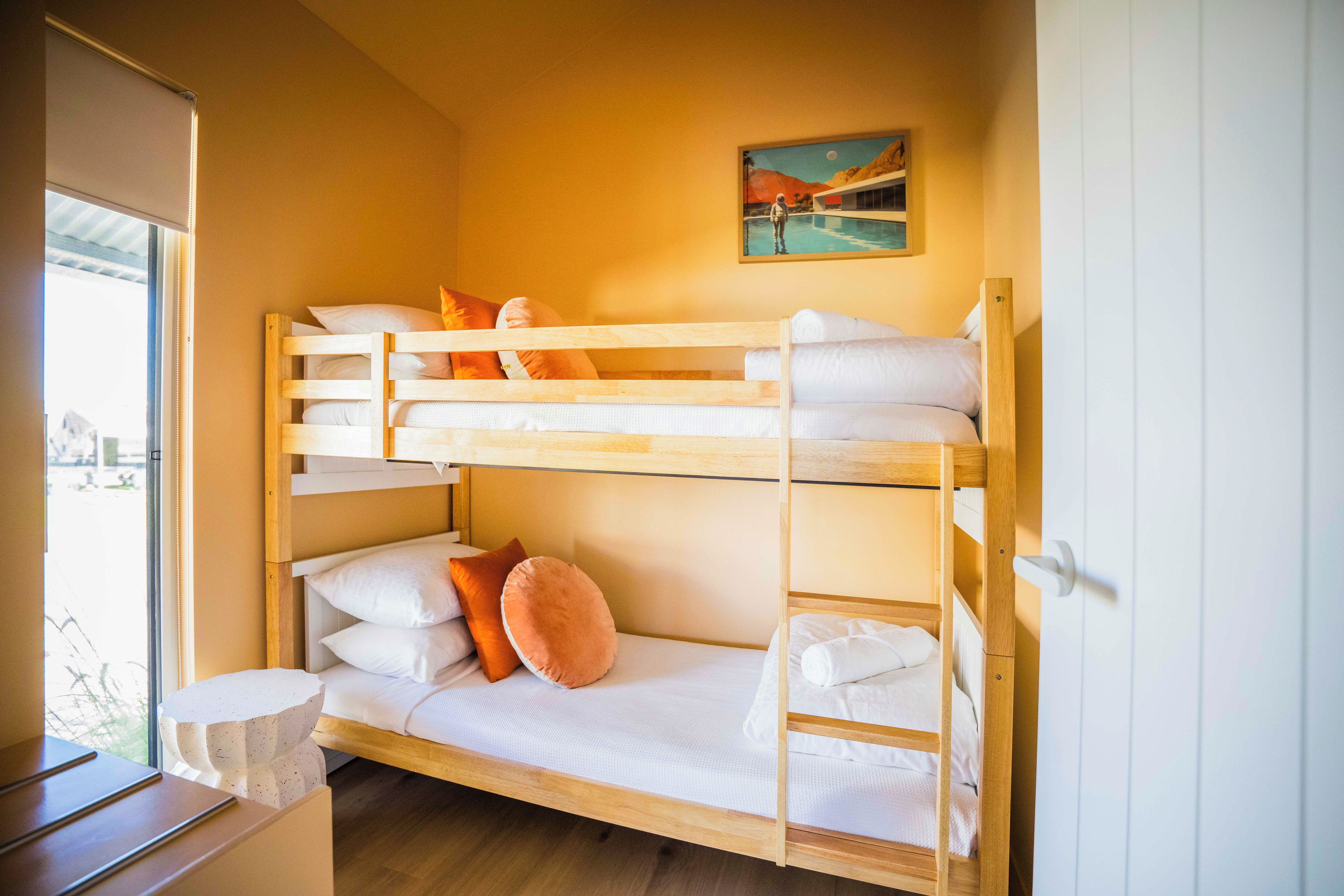 A bunk bed with white bedding and orange pillows, a painting on the wall, and a window with blinds.