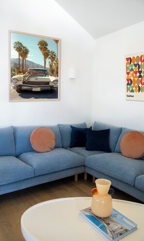 A living room with a blue sectional sofa, a round coffee table with a vase and magazine, a framed picture of a car with a leopard, and a colorful abstract art piece. - Trentham Waters Resort (Trentham Cliffs)