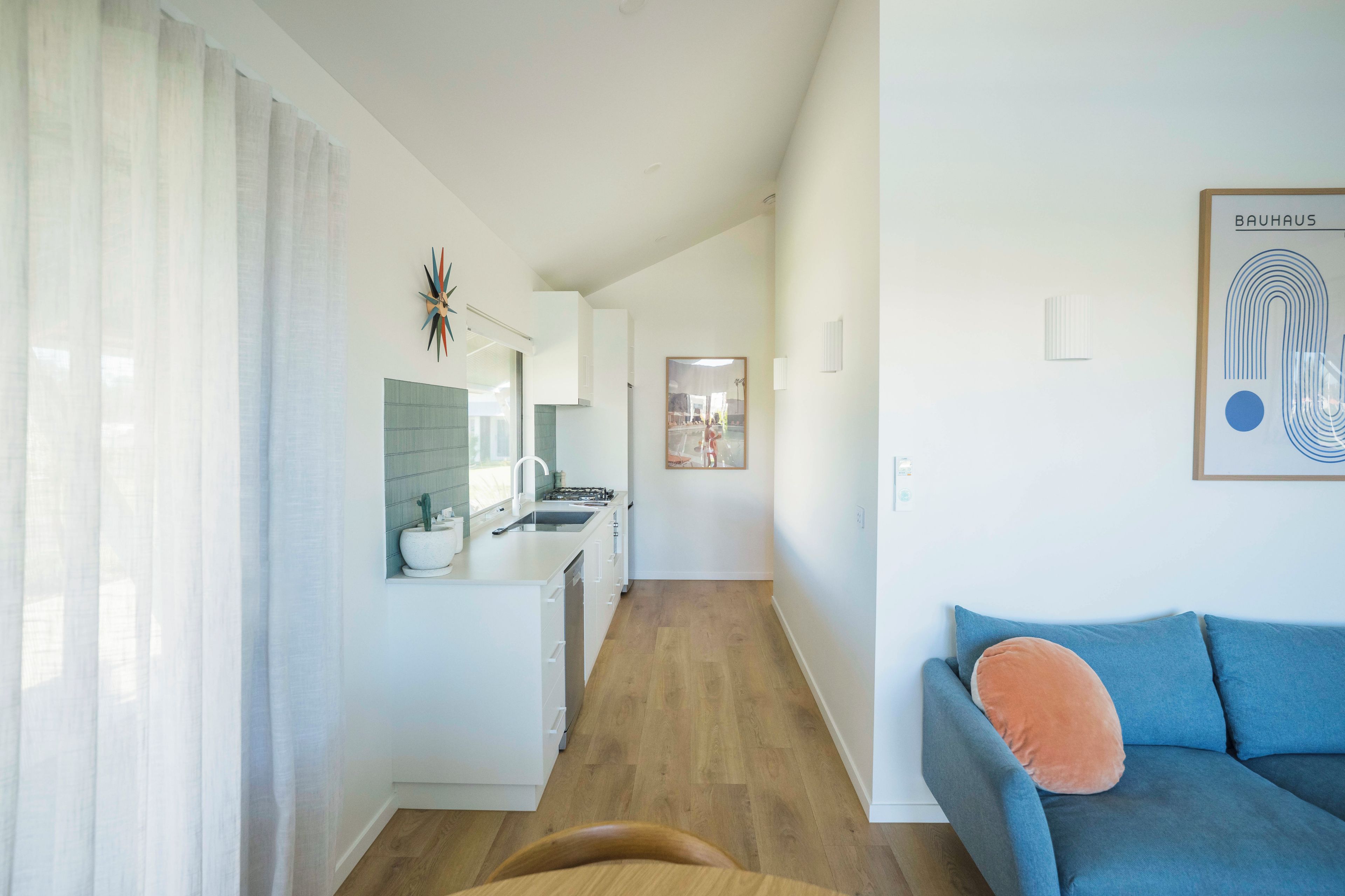A modern living room with a sofa and a coffee table, a kitchen with a sink and cabinets, and a hallway leading to another room.
