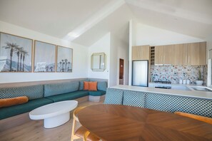 A modern living room with a green sofa, a wooden dining table, and a kitchen area with wooden cabinets and a white countertop.