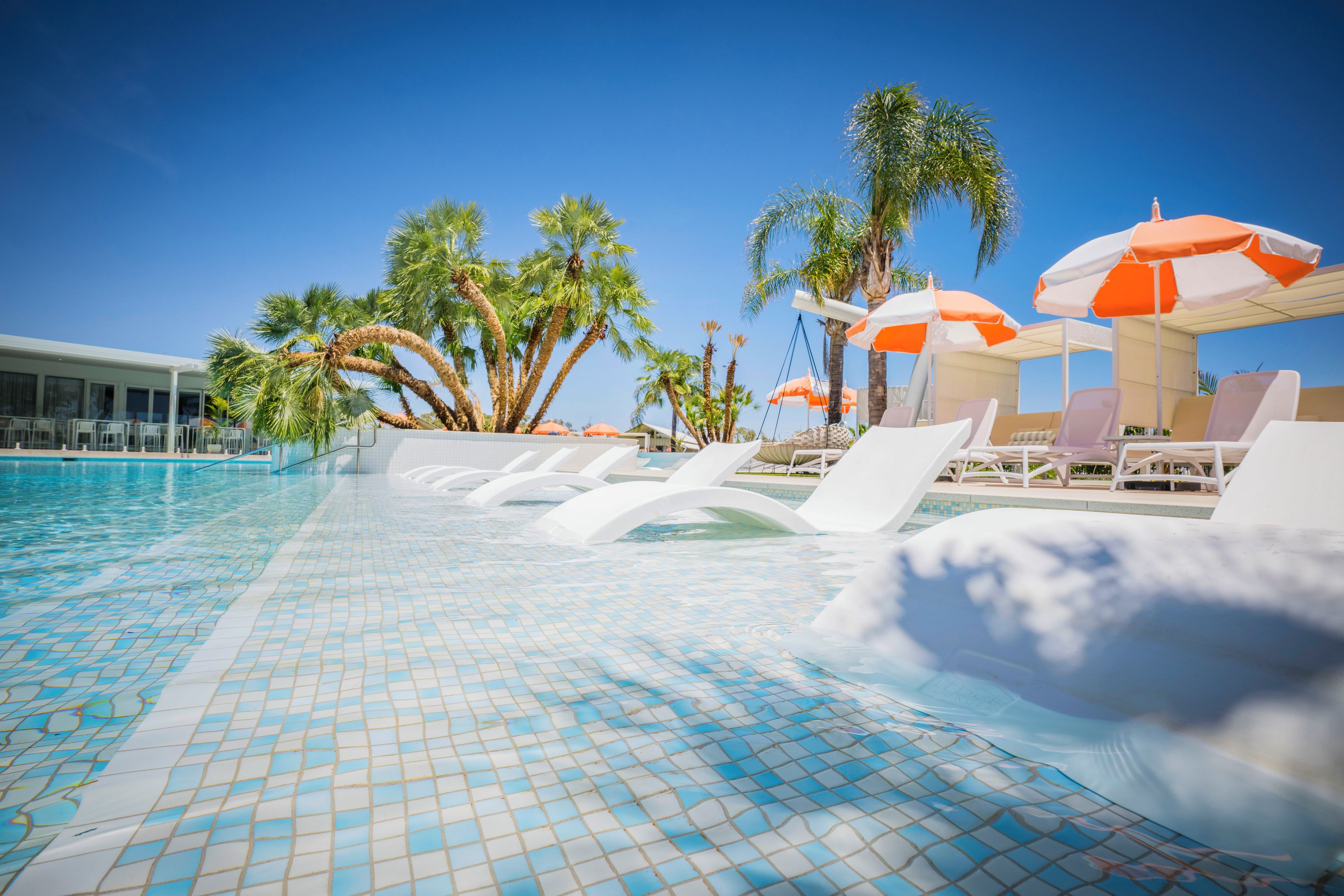 A swimming pool with tiled flooring, lounge chairs, and palm trees.