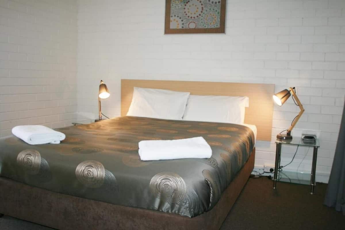 Una cama bien hecha con una colcha estampada, dos lámparas de noche, un almohadón blanco y una mesita con una lámpara.