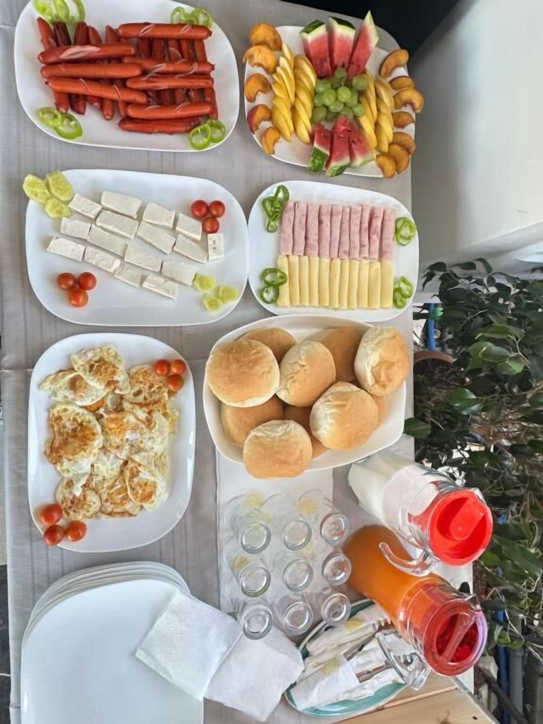 Una mesa con diversos alimentos, como perritos calientes, frutas, queso y panecillos.