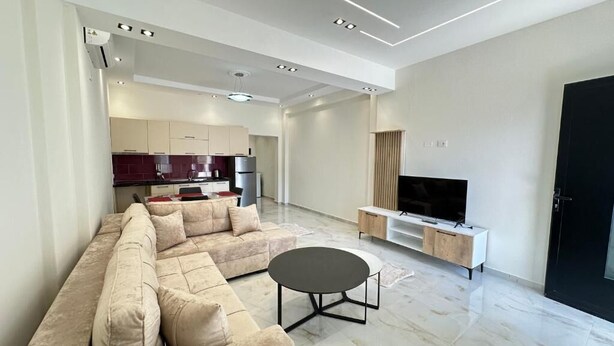 A modern living room with a sectional sofa, a round coffee table, a TV stand, and a kitchen in the background. - Liel Holiday (Vlore)