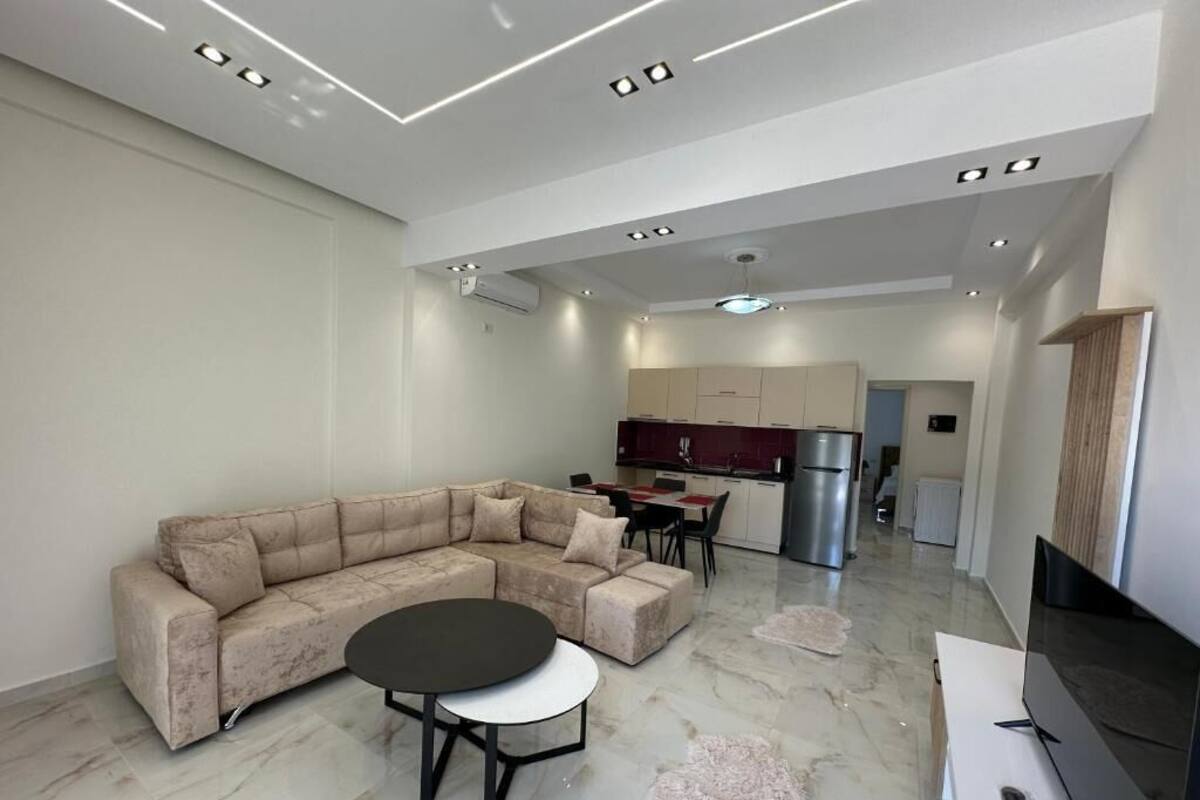 A modern living room with a sofa, a round coffee table, a flat-screen TV, and a kitchen in the background.