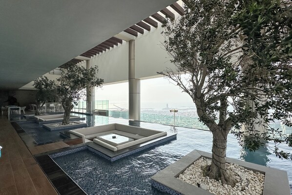 A modern indoor pool area with a view of the city, featuring a large tree, seating areas, and a water feature. - Chic 2BR in Paramount Midtown  (Dubai)