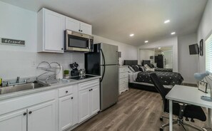 Cocina compacta con armarios blancos, refrigerador de acero inoxidable, microondas y un lavamanos. Cuenta con un pequeño comedor con una mesa y sillas.