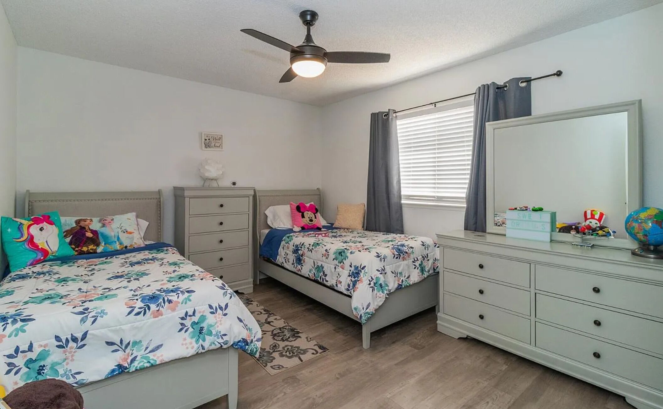 A bedroom with two beds, a ceiling fan, a dresser, a mirror, and a window with curtains.