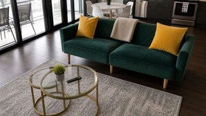 Un sofá verde con cojines amarillos, una mesa de centro de cristal con una planta y un mando a distancia encima.