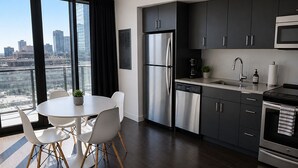 Una cocina moderna con electrodomésticos de acero inoxidable, un comedor con sillas blancas y vistas a la ciudad a través de amplias ventanas.