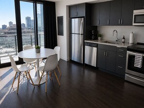 Eine moderne Küche mit Edelstahlgeräten, ein Essbereich mit weißen Stühlen und Blick auf die Stadt durch große Fenster.