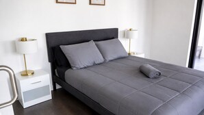 Un dormitorio moderno con una cama gris, mesitas de noche blancas y un ventanal grande.