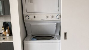 Un lavadero pequeño y blanco con lavadora y secadora de carga frontal, una pequeña encimera con utensilios de cocina y suelo de madera.