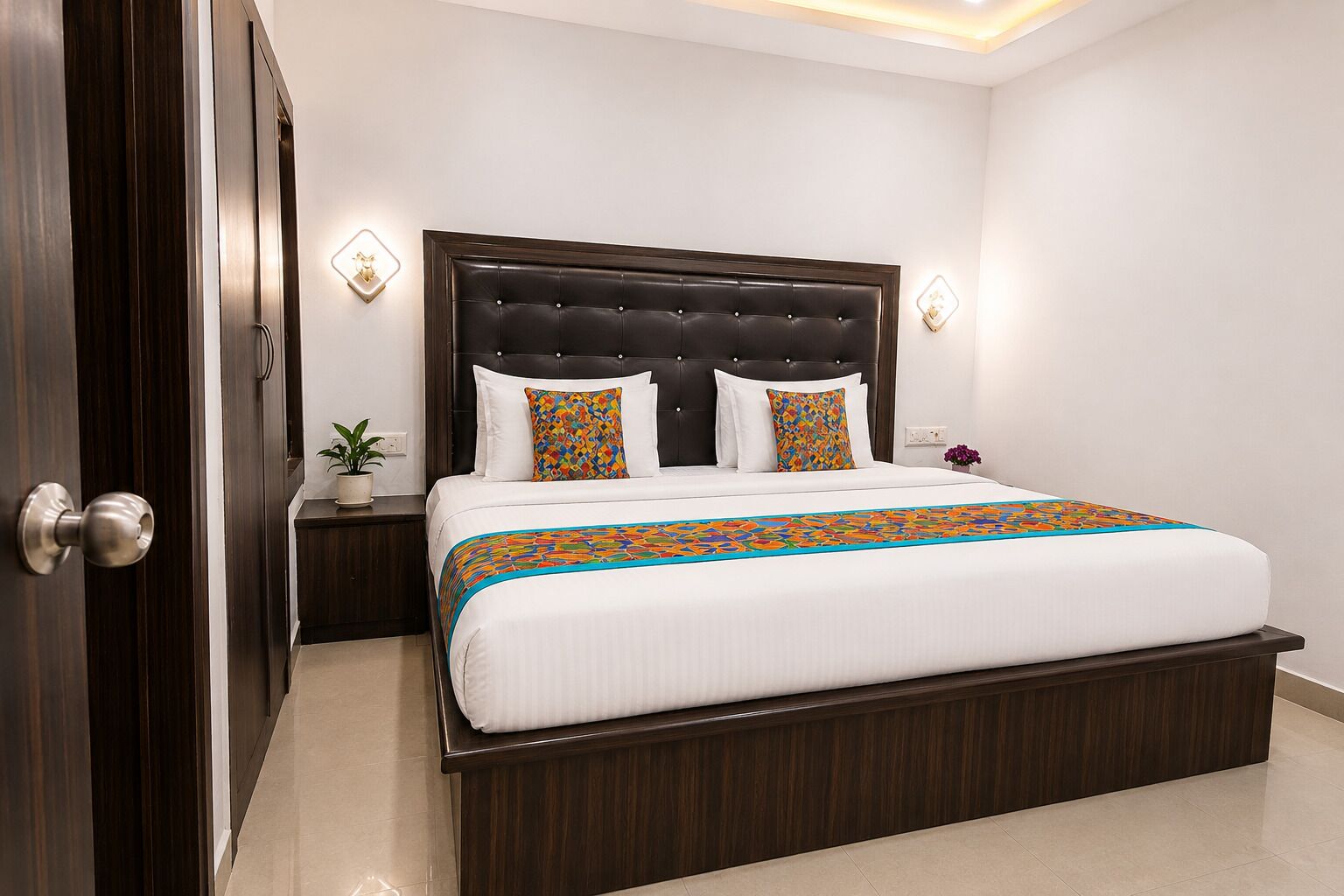 A hotel room with a large bed, dark wood headboard, white bedding, and a colorful geometric-patterned throw.