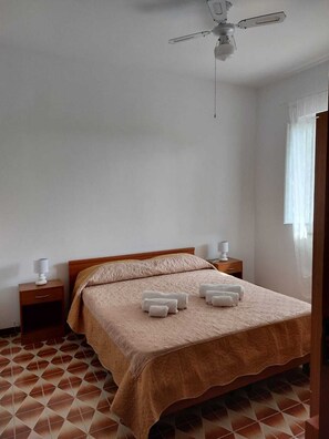 Une chambre à coucher avec un lit, deux tables de chevet, un ventilateur de plafond et une fenêtre avec des rideaux.