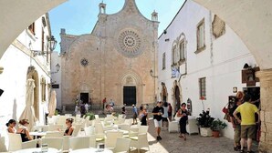 Un espace de café en plein air avec des tables et des chaises, un bâtiment historique doté d’une tour de l’horloge, et des gens qui se promènent.