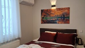 A hotel room with a bed, a painting, a radiator, a window with curtains, and a nightstand with a kettle.