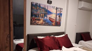 A hotel room with a bed, a wooden wardrobe, a painting of a lakeside scene, and an air conditioning unit.