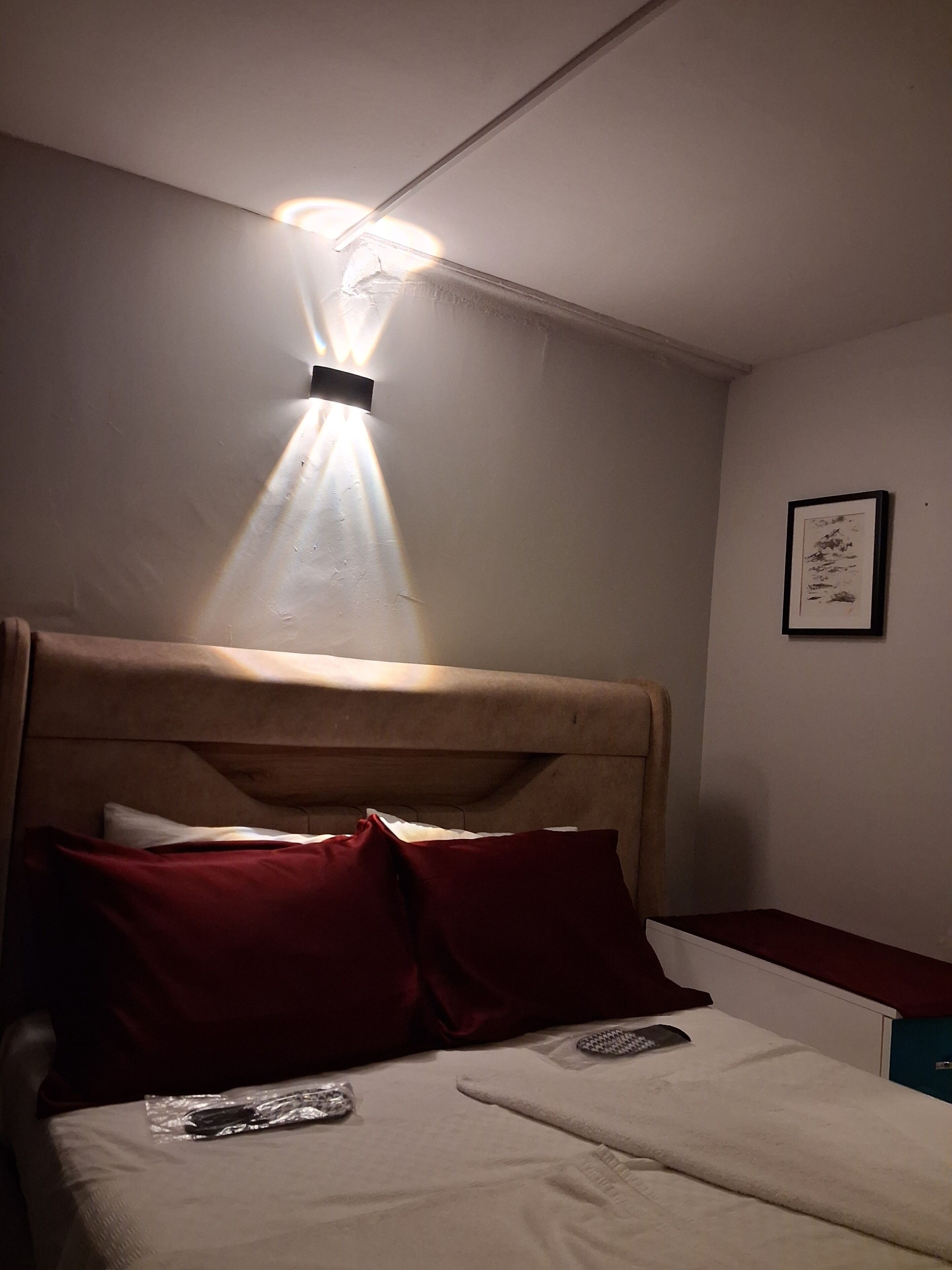Una cama con cabecera, almohadas, un control remoto y un cuadro en la pared.