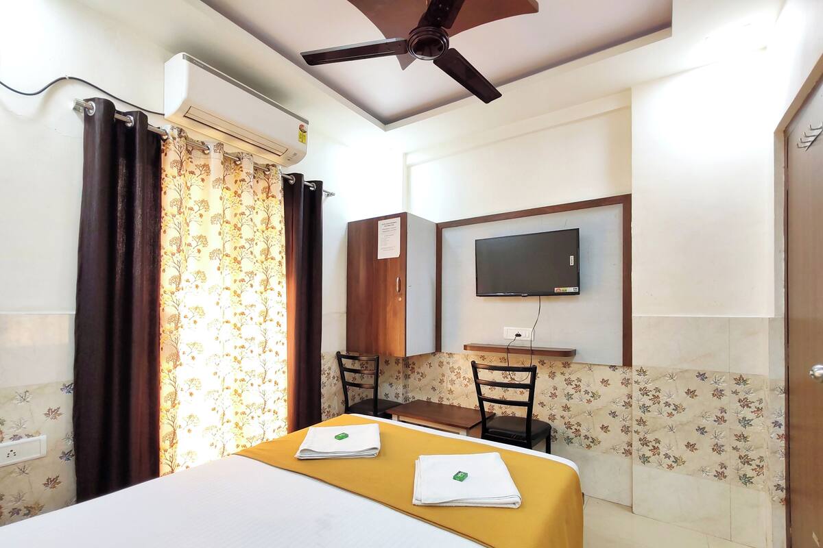 Une chambre d’hôtel avec un lit, un bureau avec une télévision, un ventilateur de plafond et une fenêtre avec des rideaux.