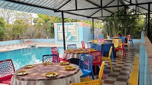 Un espacio exterior cubierto para comer con mesas y sillas, una piscina y vistas al follaje circundante.