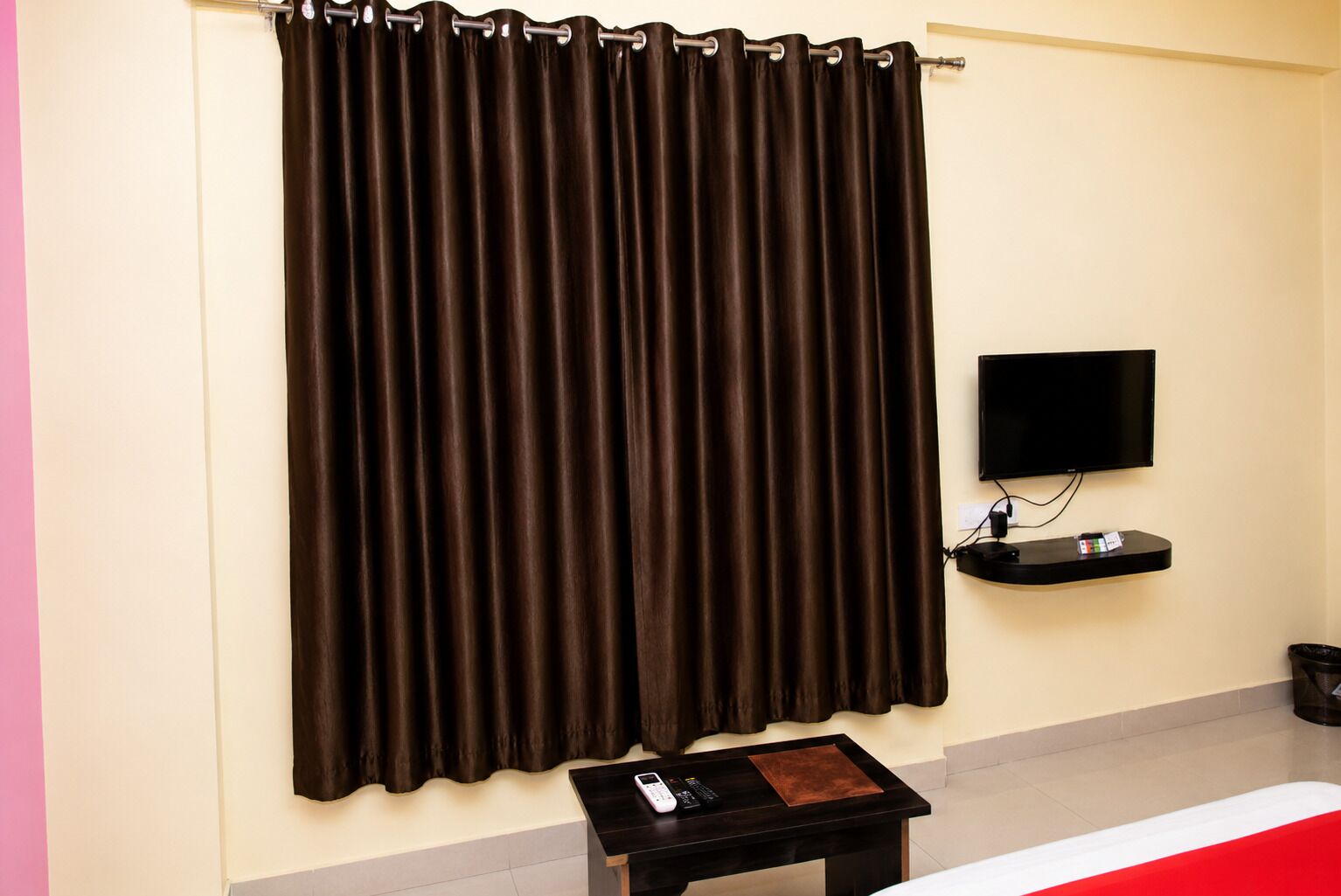 Una habitación con una cortina oscura grande, un televisor de pantalla plana montado en la pared, una mesita con un control remoto y un objeto blanco y rojo a rayas en el suelo.