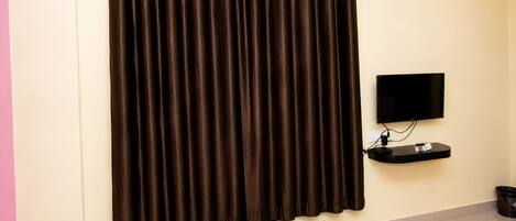 Una habitación con una cortina oscura grande, un televisor de pantalla plana montado en la pared, una mesita con un control remoto y un objeto blanco y rojo a rayas en el suelo.
