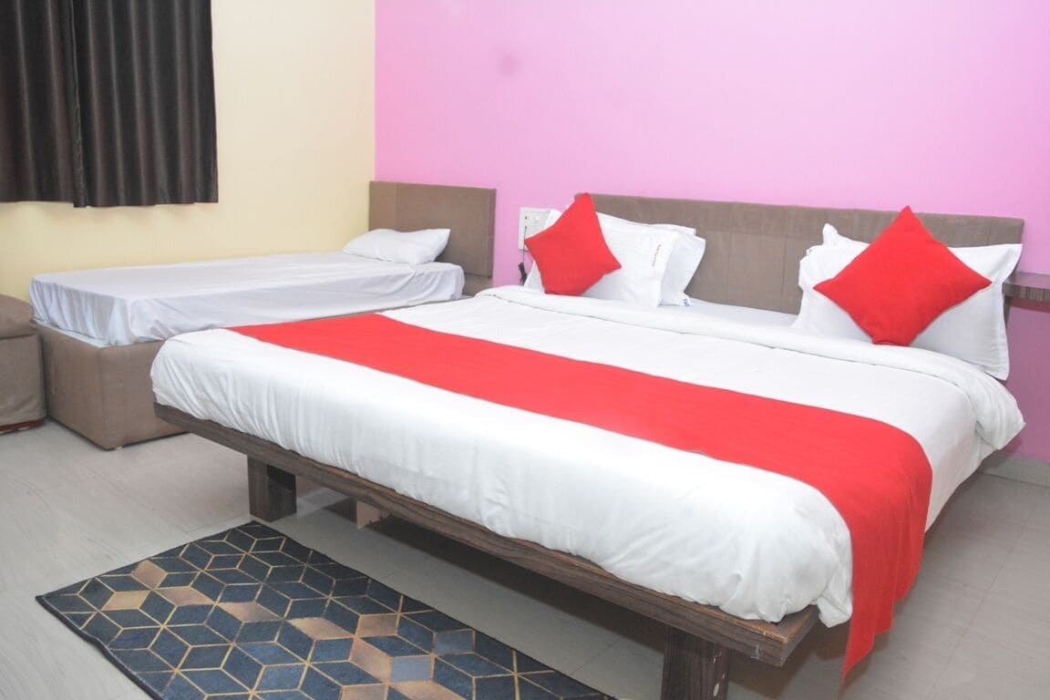 Una cama doble con una colcha roja y blanca, dos camas individuales, un cabecero marrón, una alfombra estampada en azul y negro, y una ventana con cortinas negras.