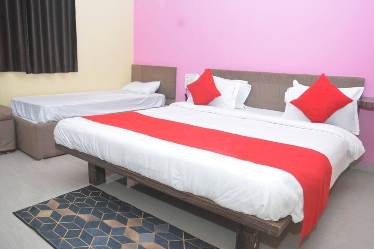 Un lit double avec une couvre-lit rouge et blanche, deux lits simples, une tête de lit marron, un tapis à motifs noirs et bleus, et une fenêtre avec des rideaux noirs.
