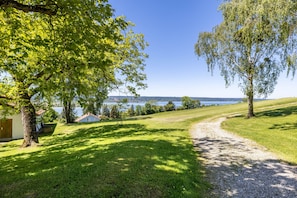 A grassy area with a gravel path leading to a body of water, surrounded by trees and a building with a red roof. - Apartment 'Hof am Ammersee' with Lake View, Balcony, and Wi-Fi (Pähl)