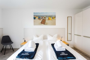 Ein Hotelzimmer mit einem Bett, zwei Nachttischen, einem Sessel, einem Strandbild und einem Kleiderschrank.