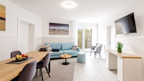 A modern living room with a sofa, a dining table with chairs, a TV mounted on the wall, and a balcony with a view. - MEERholung Vacation Rental 1.7: Close to the Beach, with Two Bedrooms and a Sofa Bed - House (Cuxhaven)