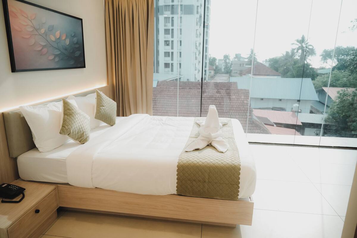 A modern hotel room with a large bed, a bedside table with a phone, a window with a view of buildings and trees, and a framed artwork on the wall.