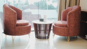 A room with two red armchairs, a small round table, and a large window with a view of buildings and trees. - Hotel Calicut Beach Annex (Kozhikode)