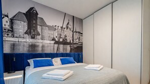 A modern bedroom with a bed, blue headboard, and a cityscape mural on the wall. - RentPlanet - Apartament przy Stoczni (Gdańsk)