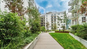 A pathway leading to a modern building with balconies, surrounded by greenery and a clear sky. - RentPlanet - Apartament przy Stoczni (Gdańsk)