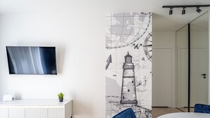 A modern room with a flat-screen TV, a white console, a potted plant, a blue chair, and a wall with a lighthouse mural. - RentPlanet - Apartament przy Stoczni (Gdańsk)
