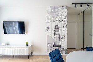 Ett modernt rum med en platt-TV, en vit konsol, en krukväxt, en blå stol och en vägg prydd med en väggmålning som föreställer ett fyrtorn.