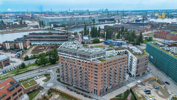 A multi-story building with a rooftop garden, surrounded by a river, industrial structures, and a mix of residential and commercial buildings. - RentPlanet - Apartament przy Stoczni (Gdańsk)