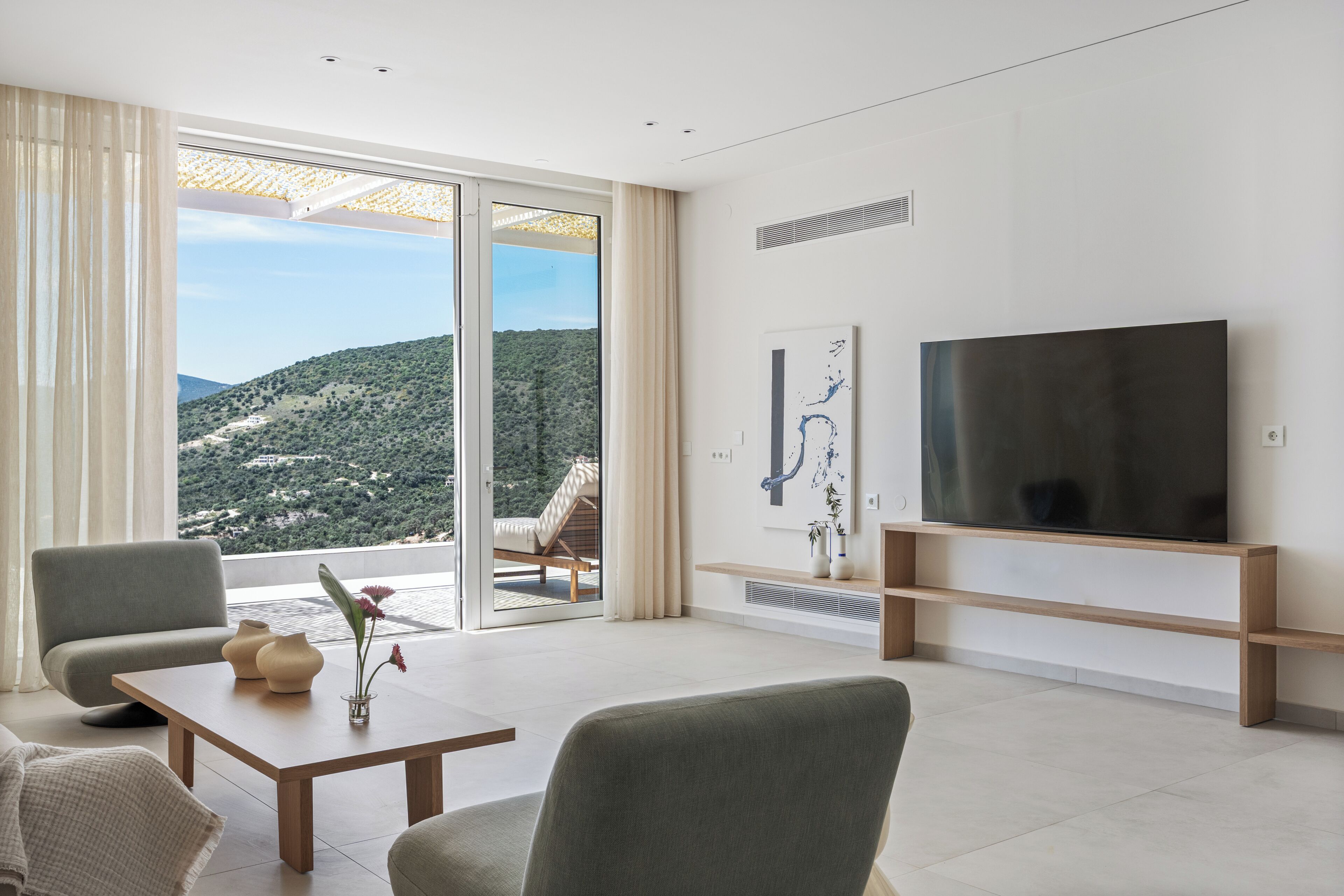 Ein modernes Wohnzimmer mit großem Fernseher, bequemen Sitzgelegenheiten, einem Couchtisch und Blick auf eine Hügellandschaft.