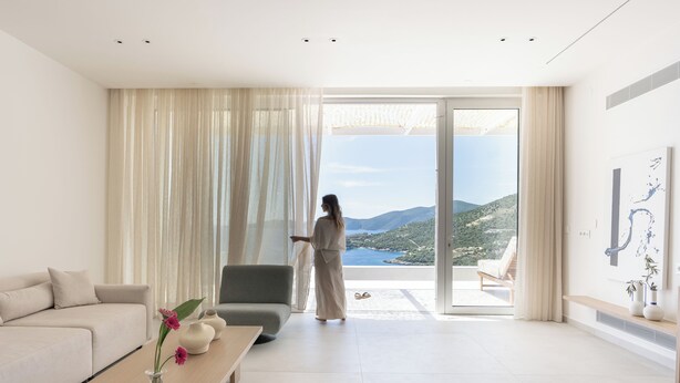 A modern living room with a sofa, a coffee table with vases, a chair, and a large sliding glass door leading to a balcony with a view of the sea and hills. - Villa Mia Rose Ammoussa Lefkada (Lefkada)